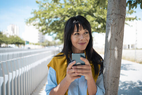 Smiling businesswoman with bangs holding smart phone leaning on tree - JOSEF19704