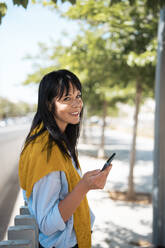 Smiling businesswoman holding smart phone near fence - JOSEF19710