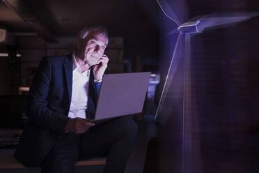 Geschäftsmann mit Laptop, der eine futuristische Windkraftanlage im Büro betrachtet - UUF28763