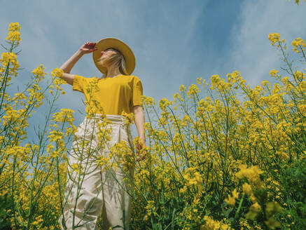 Frau mit Hut steht in einem Rapsfeld an einem sonnigen Tag - VSNF00923