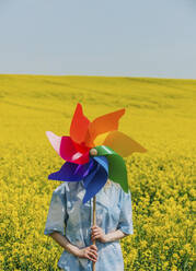 Woman holding pinwheel toy in front of face at rapeseed field - VSNF00928