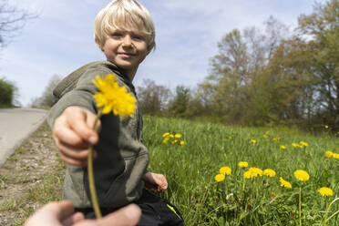 Lächelnder blonder Junge schenkt seiner Mutter auf einer Wiese eine Blume - NJAF00345