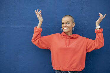 Smiling woman with shaved head dancing in front of blue wall - JCCMF10391