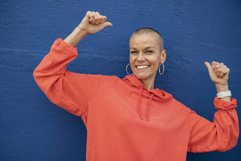 Happy woman with shaved head dancing in front of blue wall - JCCMF10392