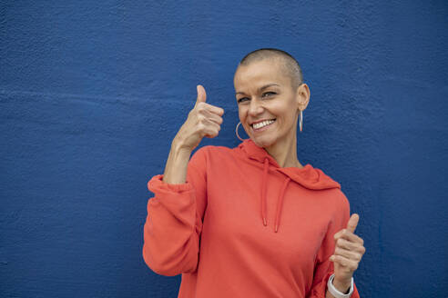 Woman with shaved head showing thumbs up in front of blue wall - JCCMF10394