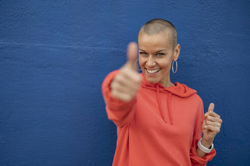 Smiling woman with shaved head showing thumbs up in front of blue wall - JCCMF10395