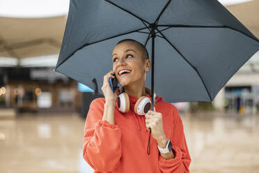 Happy shaved head woman talking on phone holding umbrella in city - JCCMF10407