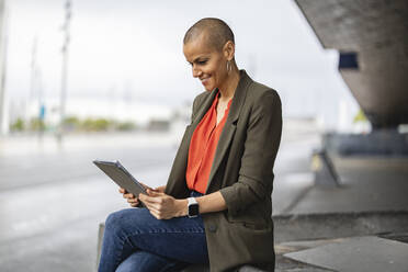 Smiling woman using tablet PC sitting on by road in city - JCCMF10413