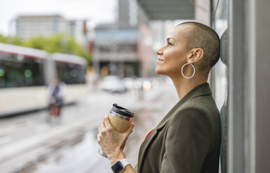 Thoughtful woman holding drink container at tram station - JCCMF10421
