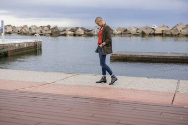 Shaved head woman with hands in pockets walking at pier - JCCMF10436