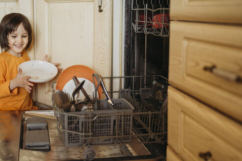 Lächelnder Junge beim Einräumen von Tellern in die Spülmaschine zu Hause - ANAF01480