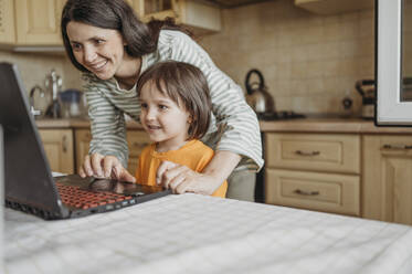 Lächelnde Mutter, die ihrem Sohn zu Hause beim Lernen mit dem Laptop hilft - ANAF01483