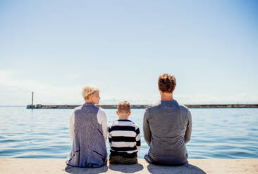 Rückansicht einer jungen Familie mit Sohn, die auf einem Steg im Freien am Meer sitzt. - HPIF20827