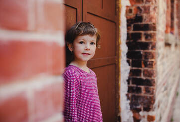 A portrait of small cute girl standing outdoors in front of door, looking at camera. - HPIF20831