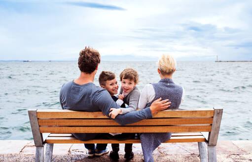 Junge Familie mit zwei kleinen Kindern sitzt auf einer Bank im Freien am Strand, Rückansicht. - HPIF20842