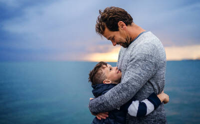 A portrait of father with small son on a walk outdoors standing on beach at dusk, hugging. - HPIF20861