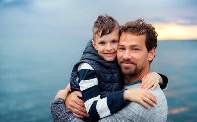 A portrait of father with small son on a walk outdoors standing on beach at dusk. - HPIF20862