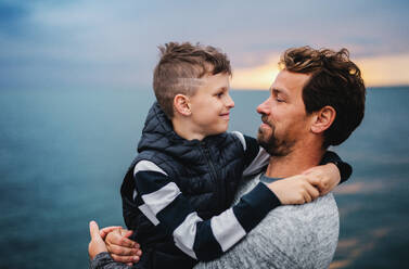 A portrait of father with small son on a walk outdoors standing on beach at dusk. - HPIF20863