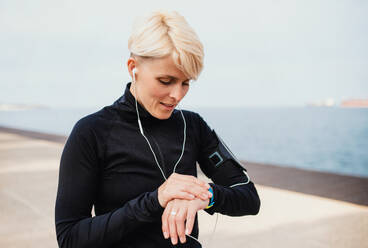 A young sportswoman with earphones standing outdoors on beach, using smartwatch. - HPIF20868