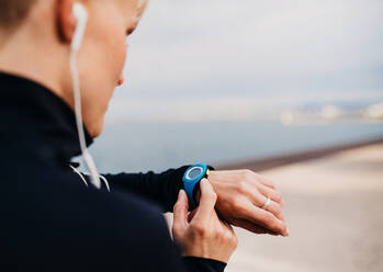 Ein Mittelteil einer jungen Sportlerin mit Kopfhörern, die draußen am Strand steht und eine Smartwatch benutzt. - HPIF20869