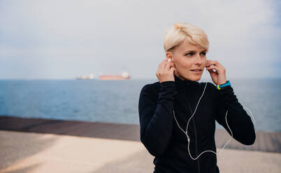 Porträt einer jungen Sportlerin mit Kopfhörern, die im Freien am Strand steht (Vorderansicht). - HPIF20870