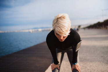 Front view portrait of young sportswoman with smatphone standing outdoors on beach, resting. - HPIF20873