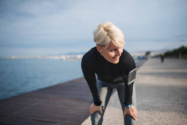 Front view portrait of young sportswoman with smartphone standing outdoors on beach, resting. - HPIF20874