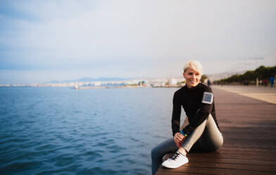 Front view portrait of young sportswoman sitting outdoors on beach, resting. Copy space. - HPIF20878