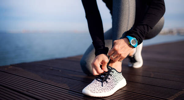 Ein Mittelteil einer jungen Sportlerin mit Smartwatch am Strand, die sich die Schnürsenkel bindet. - HPIF20888