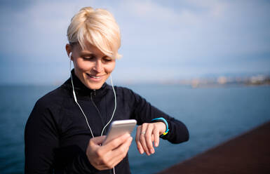 Ein Mittelteil einer jungen Sportlerin mit Kopfhörern, die im Freien am Strand steht und eine Smartwatch und ein Smartphone benutzt. - HPIF20889