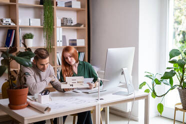 Junge Architekten mit Modell von Haus sitzen am Schreibtisch drinnen im Büro, arbeiten. - HPIF20937