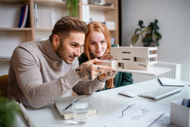 Junge Architekten mit Modell von Haus sitzen am Schreibtisch drinnen im Büro, arbeiten. - HPIF20938