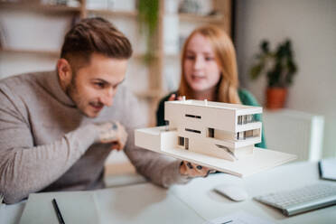 Junge Architekten mit Modell von Haus sitzen am Schreibtisch drinnen im Büro, arbeiten. - HPIF20939