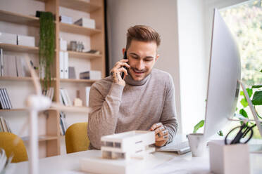 A young architect sitting at the desk indoors in office, using smartphone. - HPIF20946