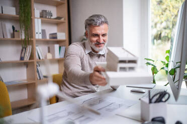 Älterer Architekt mit Hausmodell sitzt am Schreibtisch in einem Büro und arbeitet. - HPIF20961