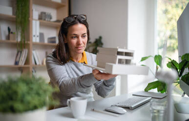 Architect with model of a house sitting at the desk indoors in office, working. - HPIF20965