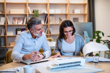 Mature architects sitting at the desk indoors in office, looking at blueprints. - HPIF20975