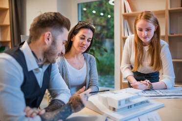 Gruppe von Architekten mit Modell von Haus sitzen am Schreibtisch drinnen im Büro, arbeiten. - HPIF20977