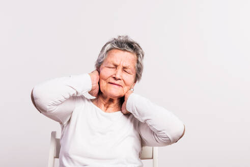Studio-Porträt einer älteren Frau mit Schmerzen, die auf einem Stuhl sitzt, weißer Hintergrund. - HPIF21080