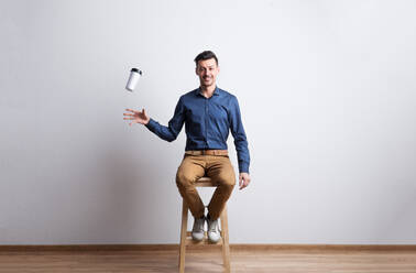 Portrait of a young handsome man in dark blue shirt sitting on a stool in a studio, trying to catch a travel coffee mug. - HPIF21094