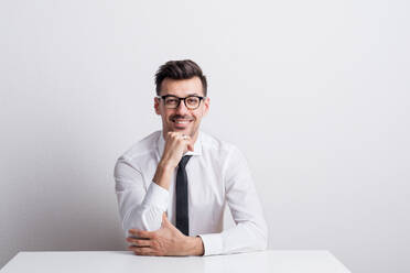 Porträt eines fröhlichen jungen Mannes mit Brille und weißem Hemd, der am Tisch in einem Atelier sitzt, die Hand am Kinn. - HPIF21100