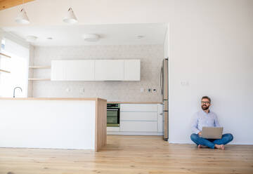 A mature man sitting on the floor in unfurnished new house, using laptop. A new home concept. - HPIF21145