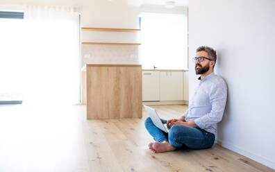 Ein reifer Mann, der in einem unmöblierten neuen Haus auf dem Boden sitzt und einen Laptop benutzt. Ein neues Hauskonzept. - HPIF21146