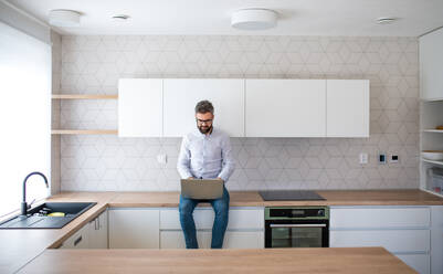 A mature man with laptop sitting in kitchen in unfurnished new house. A new home concept. - HPIF21149