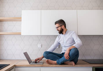 A mature man with laptop sitting in kitchen in unfurnished new house. A new home concept. - HPIF21151