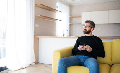 Ein glücklicher reifer Mann sitzt auf dem Sofa in einem unmöblierten Haus und hält ein Smartphone in der Hand. - HPIF21166