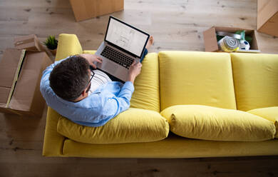 Draufsicht auf einen reifen Mann, der in einem unmöblierten Haus auf dem Sofa sitzt und einen Laptop benutzt. Ein Konzept für einen Umzug in ein neues Haus. - HPIF21204