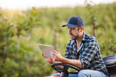 Ein seriöser, reifer Landwirt mit Tablet, der auf einem Minitraktor im Freien in einem Obstgarten sitzt. - HPIF21218