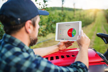 A mature farmer with tablet standing by mini tractor outdoors in orchard, checking pesticides statistics. - HPIF21219