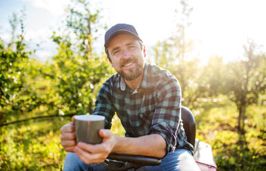 Frontansicht eines reifen Landwirts mit einer Tasse Kaffee im Freien in einem Obstgarten, der sich ausruht. - HPIF21237